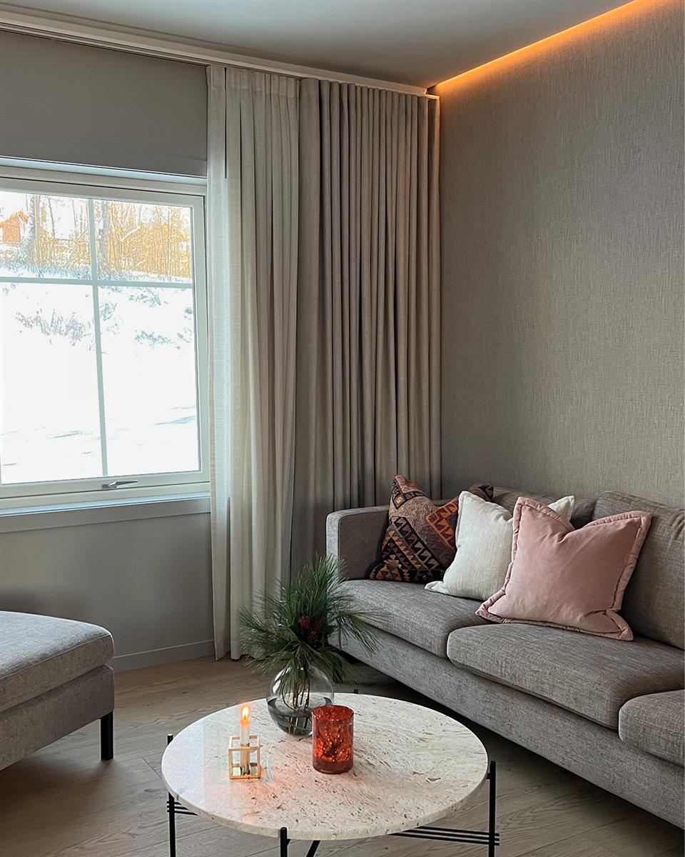 TV-stue med lyse vegger, tregulv, vindu med gardinskinne og to lag gardiner, sofa, lenestol, puff og rundt bord. Foto.